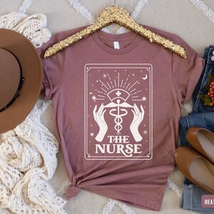 May include: A heather mauve t-shirt featuring a white graphic of a tarot card design. The card depicts a caduceus symbol with the text "THE NURSE".