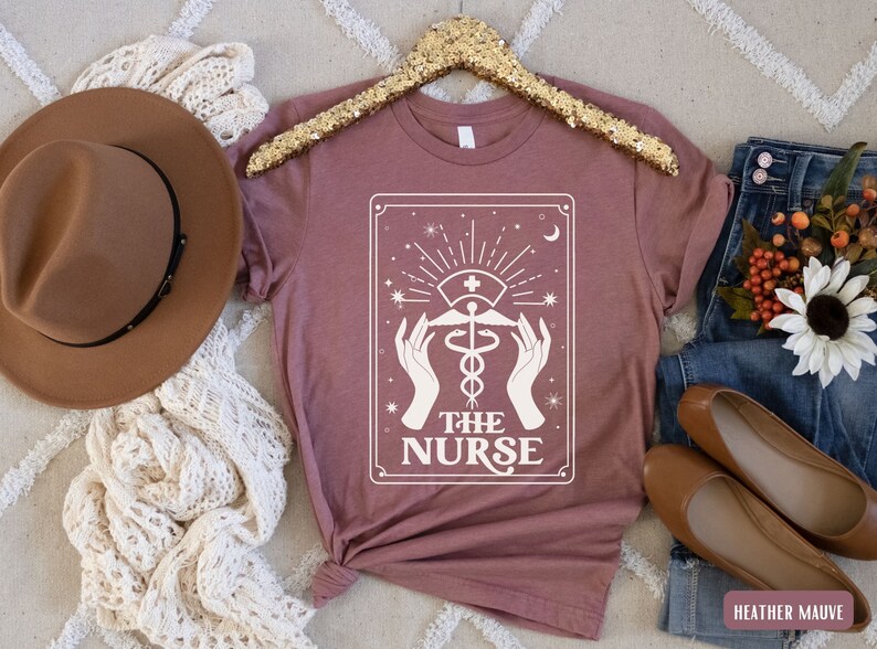 May include: A heather mauve t-shirt featuring a white graphic of a tarot card design. The card depicts a caduceus symbol with the text "THE NURSE".