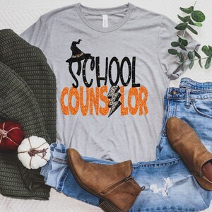 May include: A grey t-shirt with the text "School Counselor" in orange and black glitter letters. A witch's hat with a black brim and orange accents is above the text. The shirt is laid out on a white surface with a pair of brown boots, blue jeans, and a green sweater.