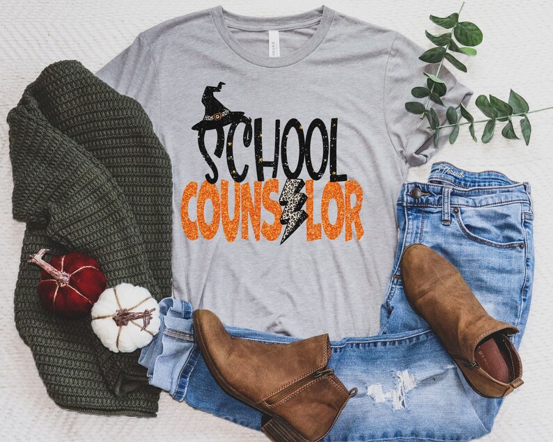 May include: A grey t-shirt with the text "School Counselor" in orange and black glitter letters. A witch's hat with a black brim and orange accents is above the text. The shirt is laid out on a white surface with a pair of brown boots, blue jeans, and a green sweater.