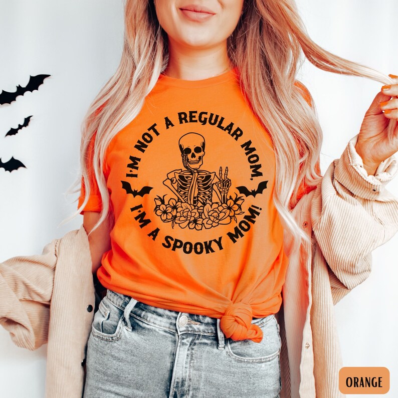 May include: Orange t-shirt with a black graphic of a skeleton sitting with flowers and bats around it. The text on the shirt reads "I'm not a regular mom. I'm a spooky mom!"