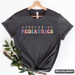 Puede incluir: Camiseta de color gris oscuro jaspeado con un gr&aacute;fico de "PEDIATRICS" de colores con flores sobre el texto.