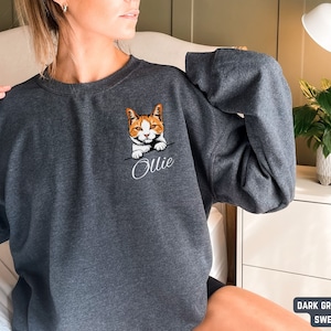 Puede incluir: Sudadera de color gris oscuro jaspeado con un gato bordado en blanco que asoma de la bolsa y el nombre "Ollie" bordado debajo.