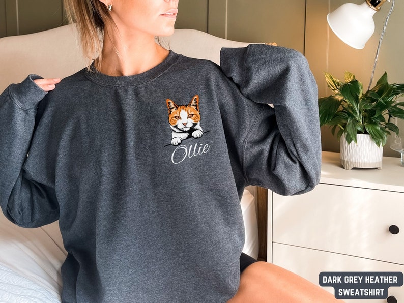 Puede incluir: Sudadera de color gris oscuro jaspeado con un gato bordado en blanco que asoma de la bolsa y el nombre "Ollie" bordado debajo.