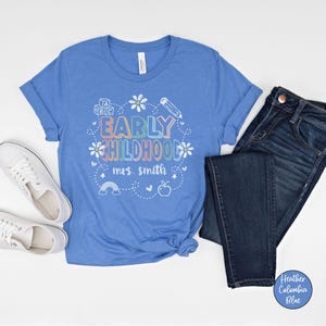 Pode incluir: Uma camiseta azul clara com as palavras "EARLY CHILDHOOD mrs. smith" em letras e gr&aacute;ficos coloridos. A camiseta &eacute; combinada com jeans azuis e t&ecirc;nis brancos. A camiseta &eacute; feita de um material macio.