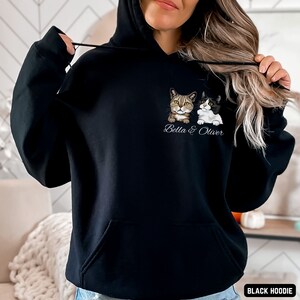 Puede incluir: Sudadera con capucha negra con un gr&aacute;fico de dos gatos, uno atigrado y otro blanco y negro, con el texto "Bella & Oliver" impreso debajo. La sudadera est&aacute; siendo modelada por una persona.