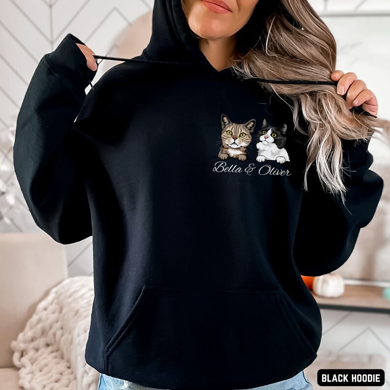 Puede incluir: Sudadera con capucha negra con un gr&aacute;fico de dos gatos, uno atigrado y otro blanco y negro, con el texto "Bella & Oliver" impreso debajo. La sudadera est&aacute; siendo modelada por una persona.