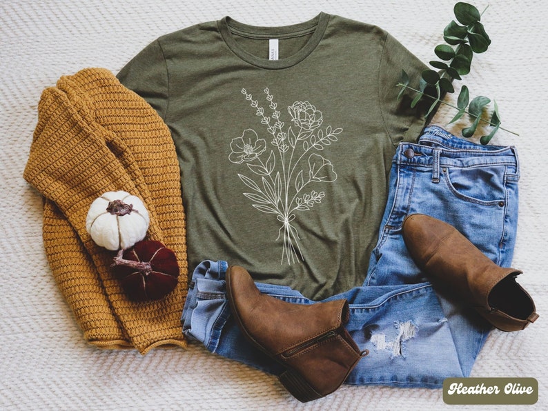 K&ouml;nnte beinhalten: Ein gr&uuml;nes T-Shirt mit einer wei&szlig;en Umrandung eines Blumenstrau&szlig;es. Das T-Shirt liegt auf einer wei&szlig;en, flauschigen Oberfl&auml;che mit einem braunen Pullover, einer blauen Jeans und braunen Stiefeln.