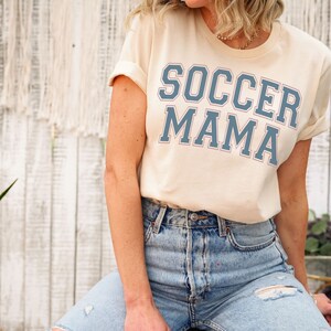 May include: A woman wearing a light beige t-shirt with the text "SOCCER MAMA" in blue lettering. She is wearing light blue jeans with a distressed look.