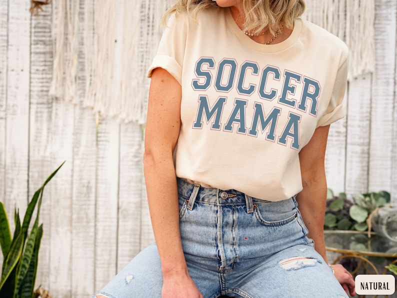 May include: A woman wearing a light beige t-shirt with the text "SOCCER MAMA" in blue lettering. She is wearing light blue jeans with a distressed look.