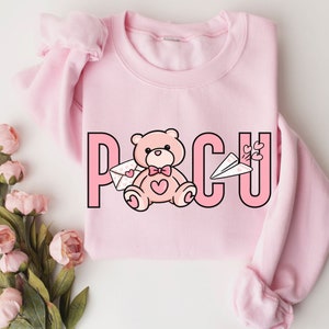 Peut inclure: Sweat-shirt rose avec un ours en peluche de dessin anim&eacute; tenant une lettre et un avion en papier. Le texte "PCU" est imprim&eacute; sur le sweat-shirt.