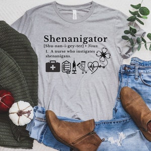 May include: A gray t-shirt with the text "Shenanigator [Shu-nan-i-gey-ter]. Noun 1. A nurse who instigates shenanigans." and a graphic of a first aid kit, a clipboard, a stethoscope, and a heart with a heartbeat line.