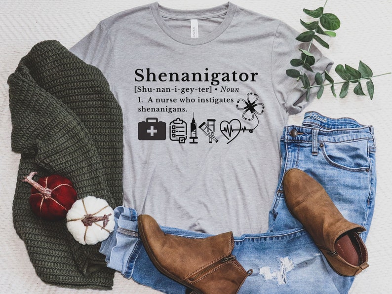 May include: A gray t-shirt with the text "Shenanigator [Shu-nan-i-gey-ter]. Noun 1. A nurse who instigates shenanigans." and a graphic of a first aid kit, a clipboard, a stethoscope, and a heart with a heartbeat line.
