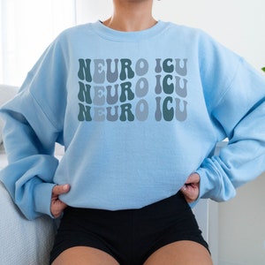 Pode incluir: Camisola azul claro com o texto "Neuro ICU" repetido tr&ecirc;s vezes em uma fonte retr&ocirc; e ondulada. O texto est&aacute; em tons de verde e azul.