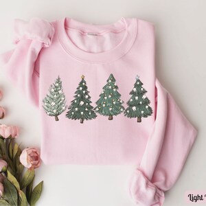 K&ouml;nnte beinhalten: Ein hellrosa Sweatshirt mit einem Motiv von vier Weihnachtsb&auml;umen, die mit Z&auml;hnen verziert sind. Die B&auml;ume sind gr&uuml;n mit wei&szlig;en und braunen Details. Das Sweatshirt hat einen Rundhalsausschnitt und lange &Auml;rmel.