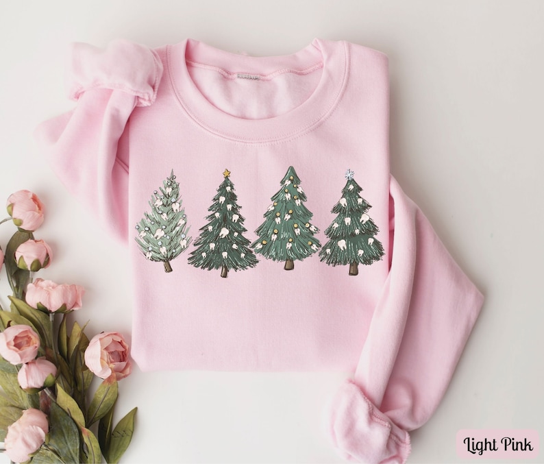 K&ouml;nnte beinhalten: Ein hellrosa Sweatshirt mit einem Motiv von vier Weihnachtsb&auml;umen, die mit Z&auml;hnen verziert sind. Die B&auml;ume sind gr&uuml;n mit wei&szlig;en und braunen Details. Das Sweatshirt hat einen Rundhalsausschnitt und lange &Auml;rmel.