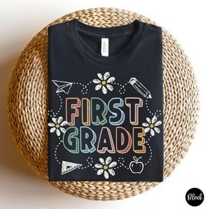 May include: Black t-shirt with the words "FIRST GRADE" in colorful letters, surrounded by white daisies, a paper airplane, a pencil, a triangle, and an apple. The shirt is displayed on a woven surface.