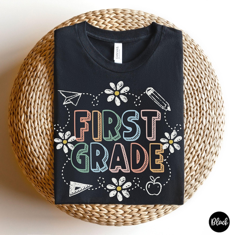 May include: Black t-shirt with the words "FIRST GRADE" in colorful letters, surrounded by white daisies, a paper airplane, a pencil, a triangle, and an apple. The shirt is displayed on a woven surface.