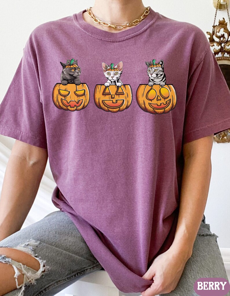 May include: A purple t-shirt with three cats wearing pumpkin hats and sitting inside pumpkins. The pumpkins have jack-o-lantern faces.