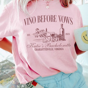 May include: Pink t-shirt with the text "VINO BEFORE VOWS" and a graphic of a vineyard, wine bottle, grapes, and cheese. The text "EST. 2024" and "Katie's Bachelorette CHARLOTTESVILLE, VIRGINIA" is also on the shirt.