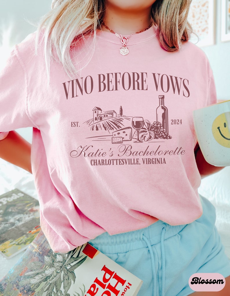 May include: Pink t-shirt with the text "VINO BEFORE VOWS" and a graphic of a vineyard, wine bottle, grapes, and cheese. The text "EST. 2024" and "Katie's Bachelorette CHARLOTTESVILLE, VIRGINIA" is also on the shirt.