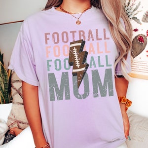 Op de afbeelding: Een lavendelkleurig T-shirt met de tekst "FOOTBALL FOOTBALL FOOTBALL MOM" in een verweerd lettertype. Een bruine voetbal met een witte bliksem bevindt zich in het midden van de tekst.