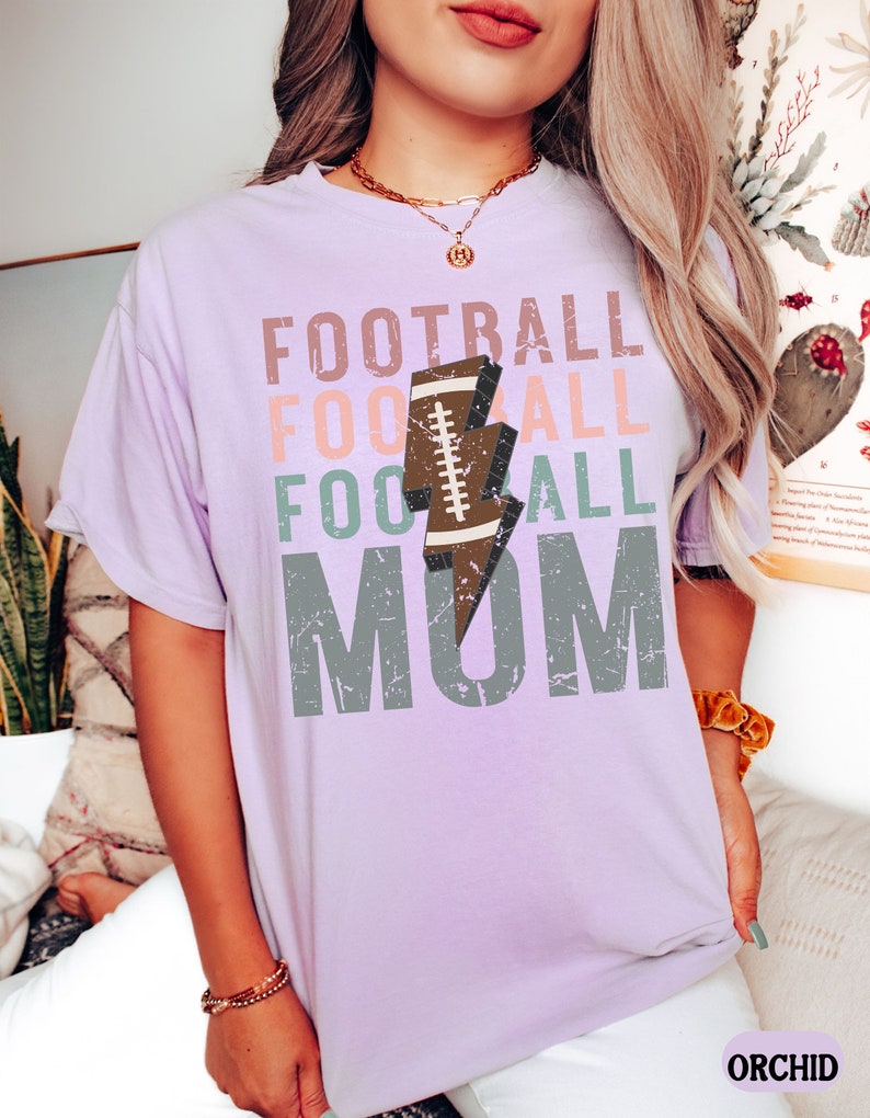 Op de afbeelding: Een lavendelkleurig T-shirt met de tekst "FOOTBALL FOOTBALL FOOTBALL MOM" in een verweerd lettertype. Een bruine voetbal met een witte bliksem bevindt zich in het midden van de tekst.