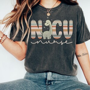 May include: A dark grey t-shirt with a striped design that spells out "NCU nurse" with a cartoon dinosaur in the middle. The dinosaur is green with a light brown belly and has a light brown neck.