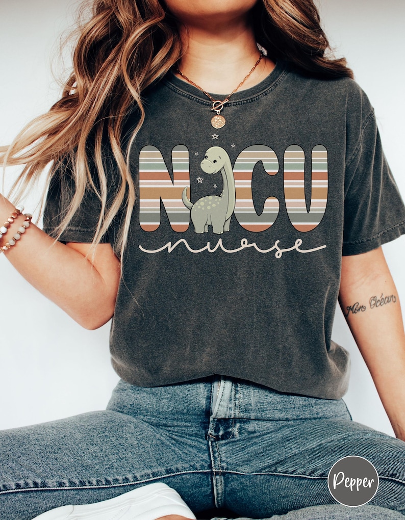 May include: A dark grey t-shirt with a striped design that spells out "NCU nurse" with a cartoon dinosaur in the middle. The dinosaur is green with a light brown belly and has a light brown neck.