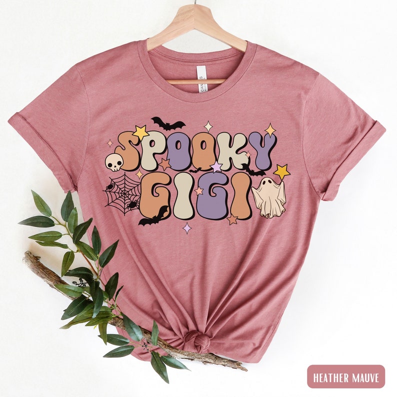 May include: Heather mauve t-shirt featuring a retro-style graphic that reads "Spooky Gigi" in colourful letters. The graphic also includes bats, stars, a spider web, a skull, and a ghost.