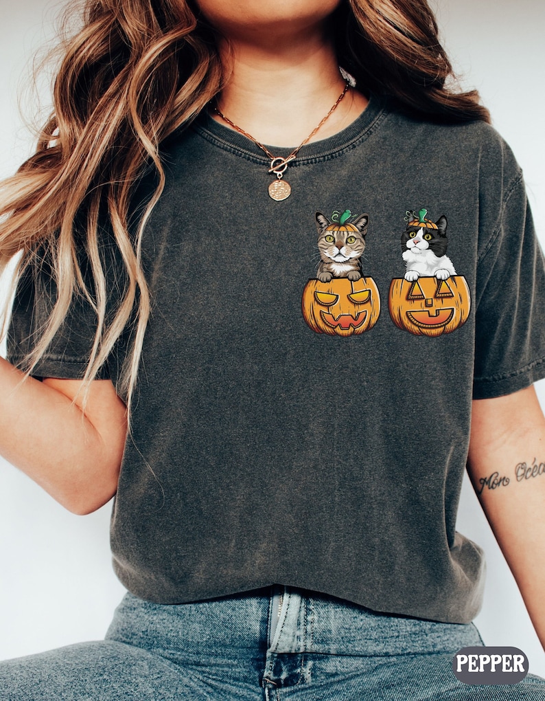 May include: A dark gray t-shirt with a graphic of two cats wearing pumpkin hats and sitting inside jack-o'-lantern pumpkins.