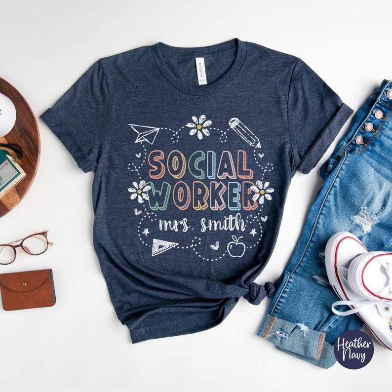 May include: Heather navy blue t-shirt with the words "SOCIAL WORKER" in colorful letters, along with the name "mrs. smith" in white script. The design includes a paper airplane, pencil, flowers, and an apple. The shirt is paired with blue jeans and white sneakers.