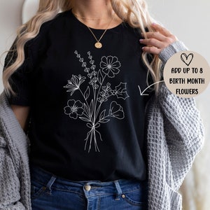 K&ouml;nnte beinhalten: Ein schwarzes T-Shirt mit einer wei&szlig;en Linienzeichnung eines Blumenstrau&szlig;es. Der Text "Addiere bis zu 8 Geburtsmonatsblumen" steht auf einem beigefarbenen Kreis mit einem Herzen.