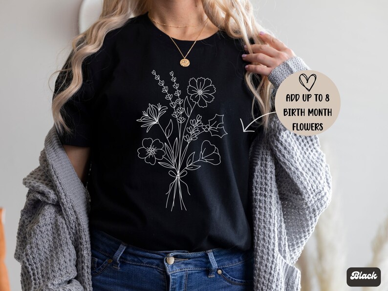 K&ouml;nnte beinhalten: Ein schwarzes T-Shirt mit einer wei&szlig;en Linienzeichnung eines Blumenstrau&szlig;es. Der Text "Addiere bis zu 8 Geburtsmonatsblumen" steht auf einem beigefarbenen Kreis mit einem Herzen.