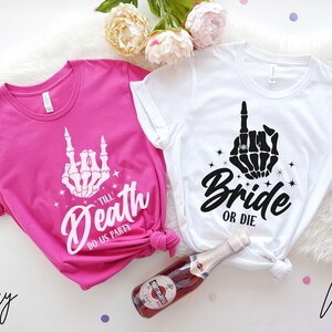 Puede incluir: Dos camisetas blancas con un dise&ntilde;o de mano de esqueleto. La camiseta rosa dice "Till Death Do Us Party" y la camiseta blanca dice "Bride or Die".