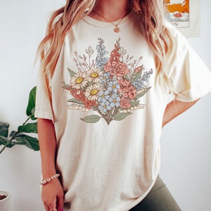 Pode incluir: T-shirt cor marfim com um design de buqu&ecirc; floral em tons de vermelho, azul e branco. O buqu&ecirc; inclui margaridas, rosas e outras flores. A camisa tem mangas curtas e um ajuste folgado. A palavra "Ivory" &eacute; impressa no canto inferior direito.