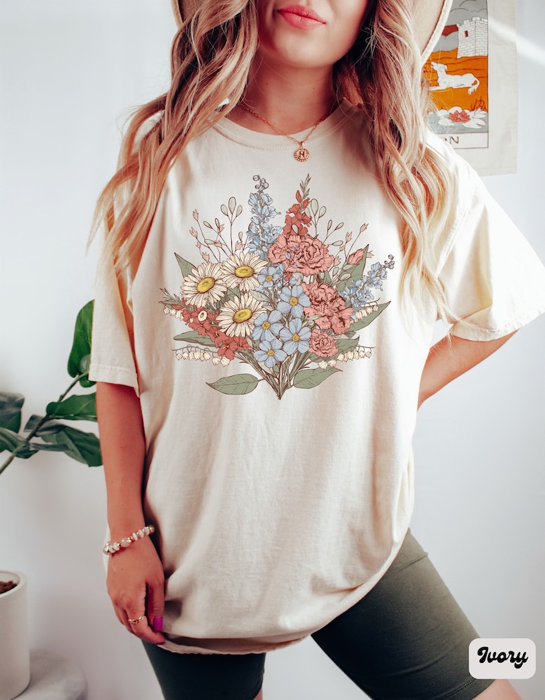 Pode incluir: T-shirt cor marfim com um design de buqu&ecirc; floral em tons de vermelho, azul e branco. O buqu&ecirc; inclui margaridas, rosas e outras flores. A camisa tem mangas curtas e um ajuste folgado. A palavra "Ivory" &eacute; impressa no canto inferior direito.