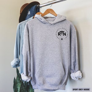 Peut inclure: Un sweat &agrave; capuche gris avec un imprim&eacute; noir qui dit "Emergency Department RN" avec un symbole m&eacute;dical. Le sweat-shirt est suspendu &agrave; un cintre.