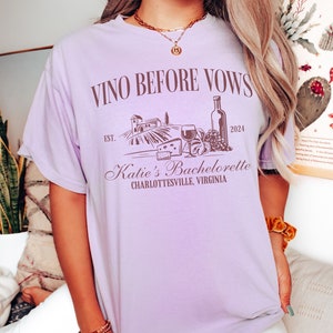 May include: Purple t-shirt with the text "Vino Before Vows" and a graphic of a vineyard, wine bottle, grapes, cheese, and a glass of wine. The text "Est. 2024" and "Katie's Bachelorette Charlottesville, Virginia" is also on the shirt.