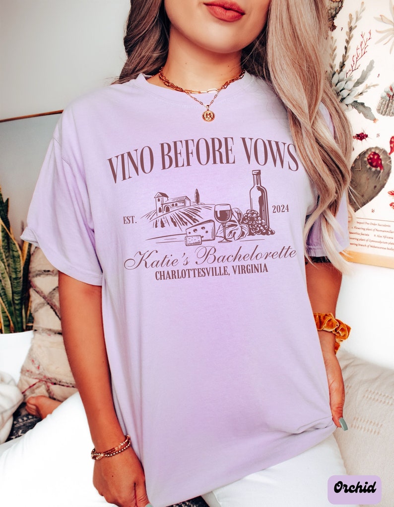 May include: Purple t-shirt with the text "Vino Before Vows" and a graphic of a vineyard, wine bottle, grapes, cheese, and a glass of wine. The text "Est. 2024" and "Katie's Bachelorette Charlottesville, Virginia" is also on the shirt.