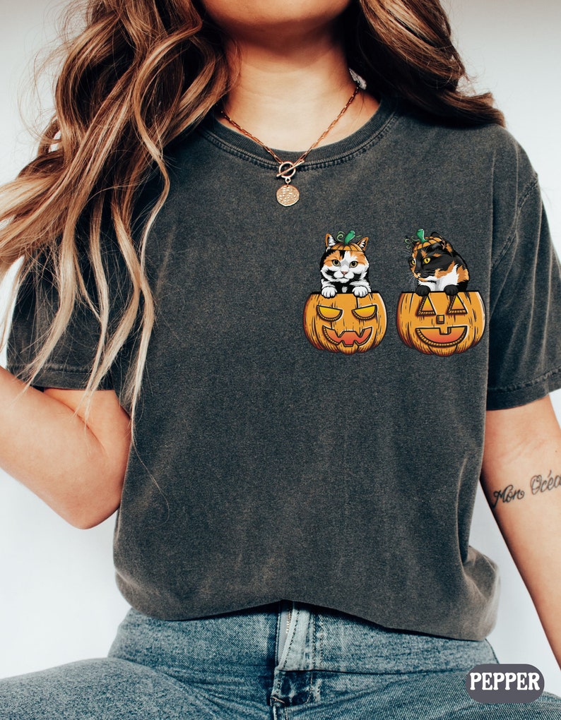 May include: A dark grey t-shirt featuring a graphic of two calico cats peeking out of jack-o'-lantern pumpkins.