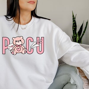 Peut inclure: Sweat-shirt blanc avec un motif rose repr&eacute;sentant un ours en peluche tenant une lettre et un avion en papier. Le motif comprend &eacute;galement le texte "PCU" en lettres roses.