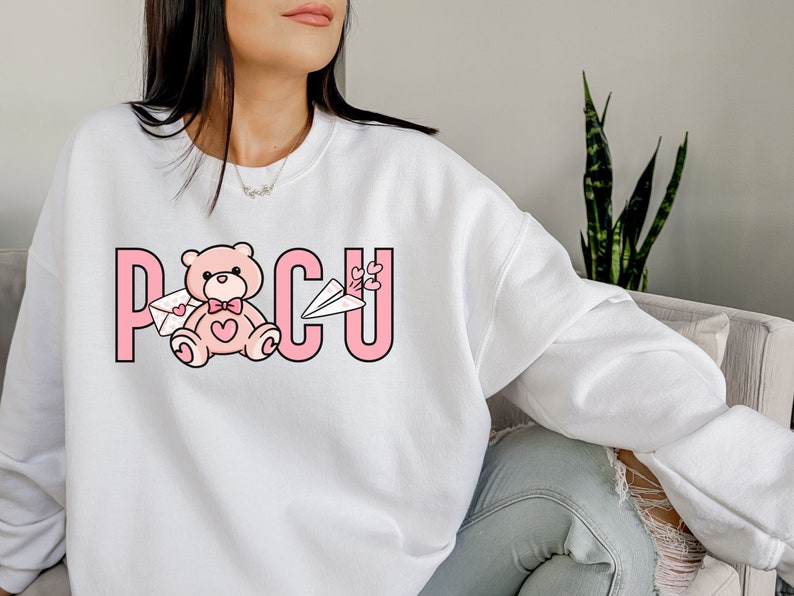 Peut inclure: Sweat-shirt blanc avec un motif rose repr&eacute;sentant un ours en peluche tenant une lettre et un avion en papier. Le motif comprend &eacute;galement le texte "PCU" en lettres roses.