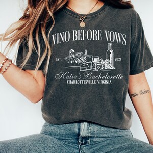 May include: A dark gray t-shirt with the text "Vino Before Vows" and a graphic of a vineyard, wine bottle, glasses, and cheese. The text "Katie's Bachelorette Charlottesville, Virginia" is also on the shirt.