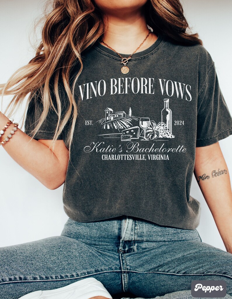 May include: A dark gray t-shirt with the text "Vino Before Vows" and a graphic of a vineyard, wine bottle, glasses, and cheese. The text "Katie's Bachelorette Charlottesville, Virginia" is also on the shirt.