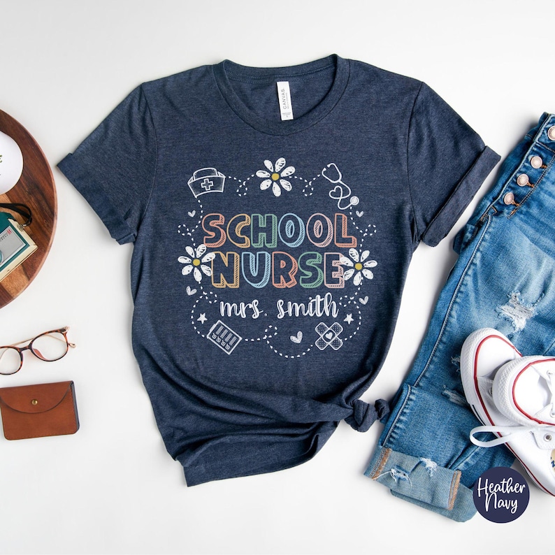 Puede incluir: Camiseta azul marino con la frase "SCHOOL NURSE mrs. smith" en un dise&ntilde;o colorido. La camiseta presenta un tema m&eacute;dico con margaritas, un estetoscopio y un gr&aacute;fico de vendaje. La camiseta se combina con vaqueros azules y zapatillas blancas.