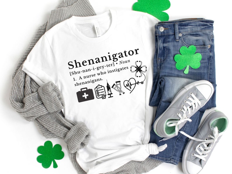May include: White t-shirt with a black graphic that says "Shenanigator [Shu-nan-i-gey-ter]. Noun 1. A nurse who instigates shenanigans."  The graphic also includes a first aid kit, a clipboard, a syringe, a stethoscope, a heart, and a four-leaf clover.