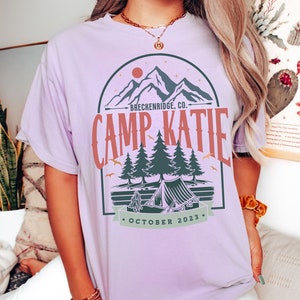 May include: A lavender t-shirt with a graphic design of a mountain range, a tent, and trees. The text on the shirt reads "Breckenridge, Co. Camp Katie October 2023."