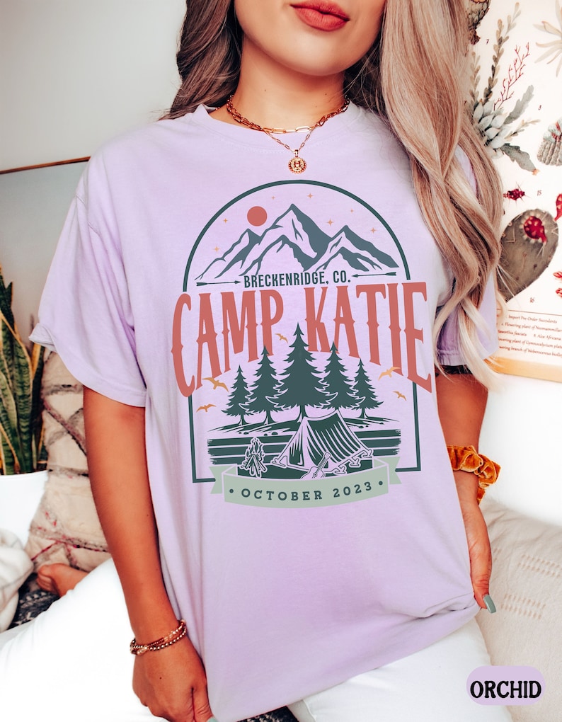 May include: A lavender t-shirt with a graphic design of a mountain range, a tent, and trees. The text on the shirt reads "Breckenridge, Co. Camp Katie October 2023."