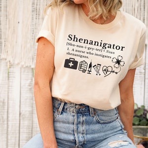 May include: A beige t-shirt with the text "Shenanigator [Shu-nan-i-gey-ter]. Noun 1. A nurse who instigates shenanigans." and a series of medical icons including a first aid kit, a clipboard, a syringe, a vial, a heart rate monitor, and a four-leaf clover.